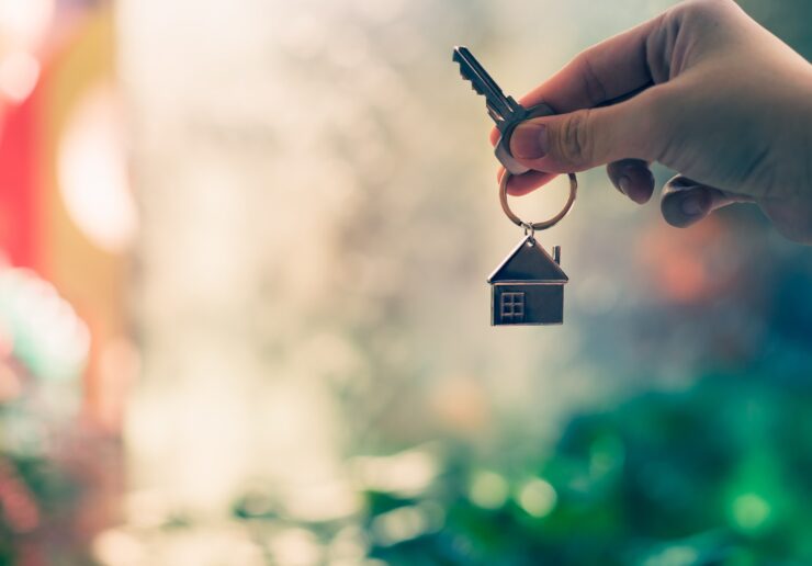 a person's right hand holding a door key on a house-shaped keyring against a blurred background.