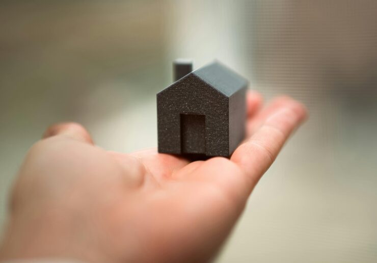 Is now a good time to buy a house or get a mortgage? Image is of an outstretched left hand with a small, dark grey, model house resting on the fingers. The background cannot be discerned, it is softly blurred.