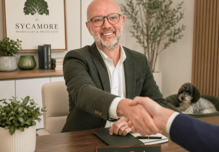 Alex Cooke, a Mortgage Broker, is sat at a desk in a light, modern office, shaking hands with an unseen client sat opposite him. In the corner behind Alex is a black and white cockerpoo dog laying in its bed.